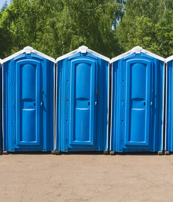 Several blue portable toilets lined up in a row against a backdrop of green trees and a clear sky.
